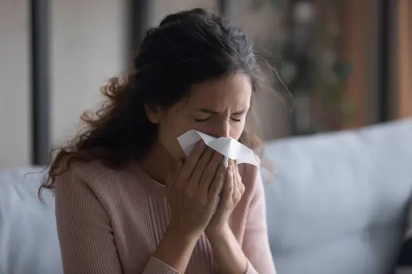 Sick young woman sitting indoors holding tissue blowing running nose — Stock Photo, Image