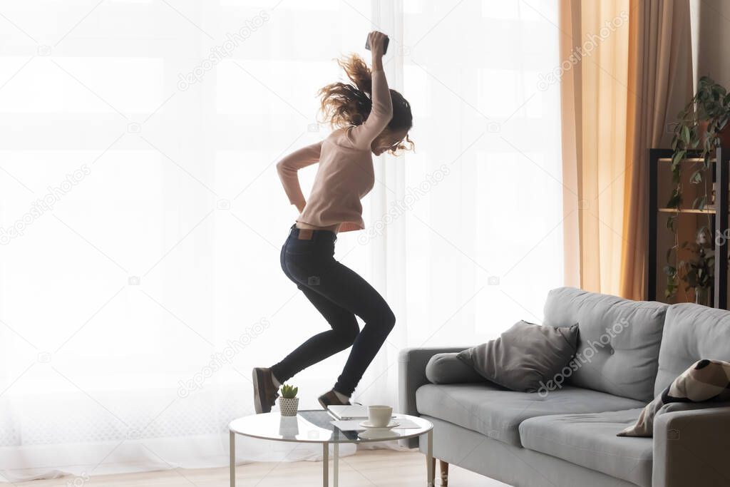 Crazy happy young woman holding cellphone jumps dances alone indoor