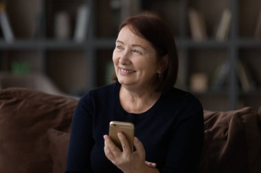 Rüya gibi gülümseyen olgun bir kadın akıllı telefonu elinde tutuyor..