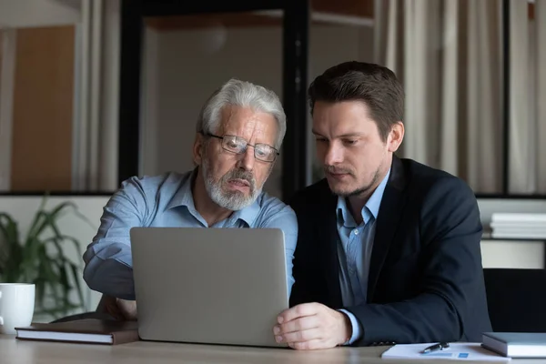 Confident mature businessman mentor training new employee, using laptop