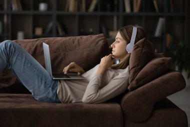 Kulaklıklı kadın dizüstü bilgisayarla kanepede yatıyor.