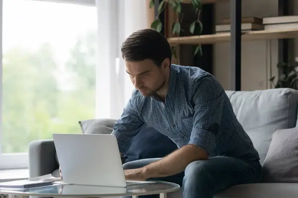 Serieuze duizendjarige man met behulp van laptop, typen, zitten op de bank — Stockfoto