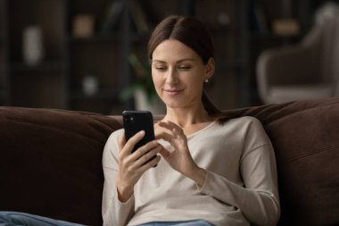 Akıllı telefon kullanan, kanepede oturan tatmin olmuş genç bir kadın.