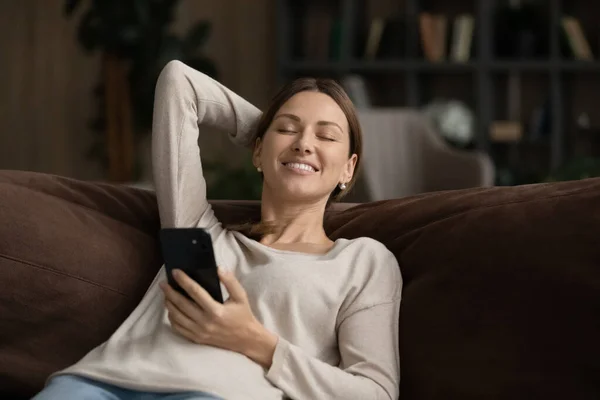 Mujer sonriente con los ojos cerrados sosteniendo teléfono inteligente, relajarse en el sofá — Foto de Stock