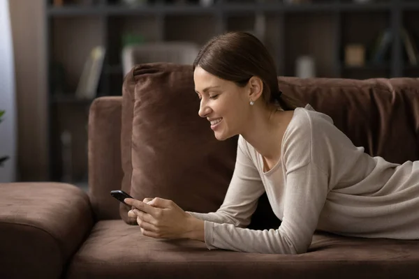 Вид сбоку улыбающаяся женщина с помощью смартфона, лежащая на уютном диване — стоковое фото