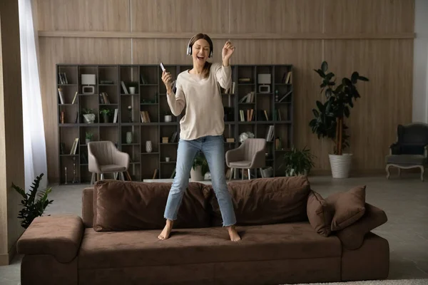 Zabawiona kobieta w słuchawkach ze smartfonem, tańcząca na kanapie — Zdjęcie stockowe