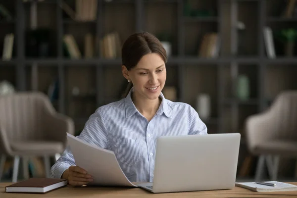 Улыбающаяся женщина с документами, работающая на ноутбуке, сидящая за столом — стоковое фото