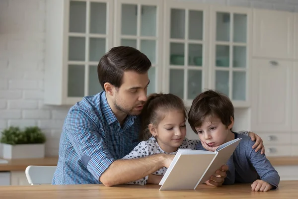 Serious millennial dad reading book to little son and daughter — Stock Photo, Image
