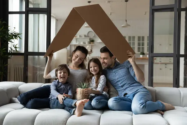 Happy sibling kids and parents holding toy roof above heads — Stock Photo, Image