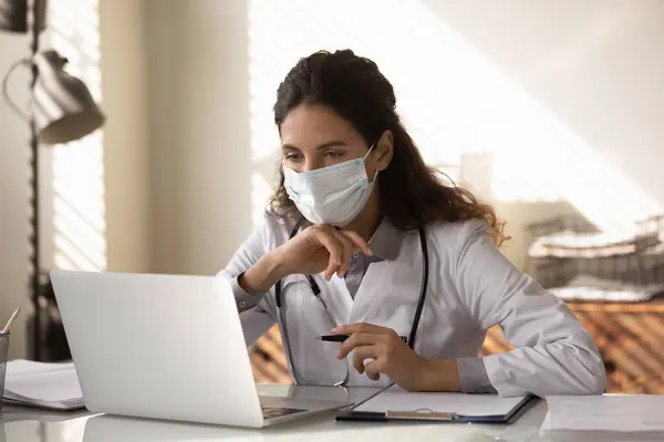 의료용 마스크를 쓰고 있는 젊은 의사가 노트북에서 일하고 있습니다. — 스톡 사진