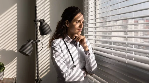 Sonriente doctora pensativa parada junto a la ventana en el consultorio — Foto de Stock