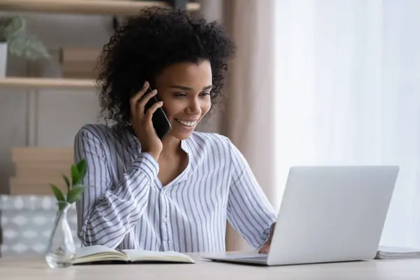Black female office employee multitasking talking by phone using laptop — Stock Photo, Image