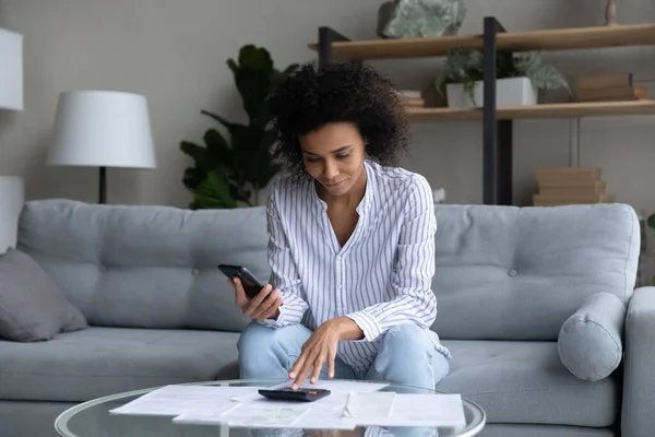 Jonge zwarte vrouw tellen op rekenmachine betalen rekeningen met behulp van telefoon — Stockfoto