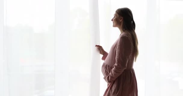 Жінка, що стоїть в приміщенні, торкається її вагітного живота, дивлячись у вікно — стокове відео