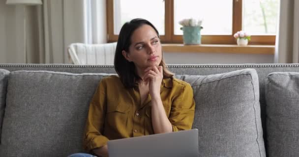 Nachdenkliche junge Frau sitzt auf Sofa und arbeitet ferngesteuert am Laptop — Stockvideo