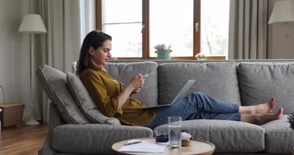 Ung kvinna som använder mobiltelefon medan du sitter på soffan med laptop — Stockvideo