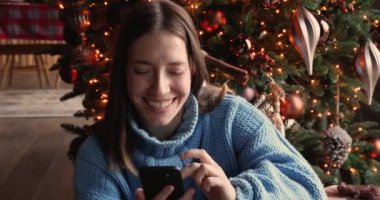 Mutlu genç kadın akıllı telefon kullanıyor, Noel ağacının yanında oturuyor..