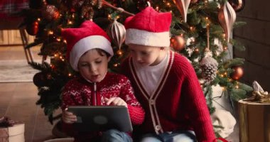 Küçük şirin çocuklar tablet kullanıyor, Noel ağacının yanında oturuyorlar..