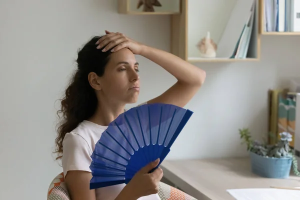 Přehřátá žena sedět uvnitř se cítí přehřáté držení modrého ventilátoru — Stock fotografie