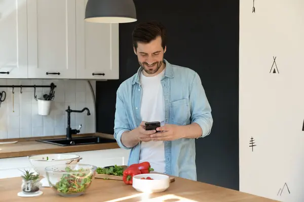 Homme cuisine dans la cuisine tenant smartphone recherche recettes en ligne — Photo