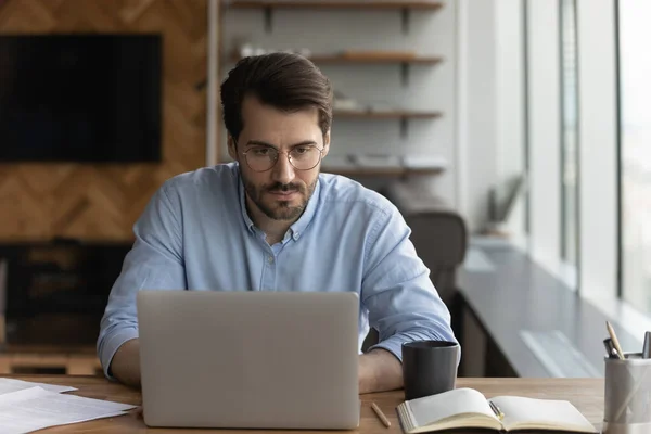 L'uomo serio guarda il lavoro dello schermo del computer portatile online sul posto di lavoro — Foto Stock