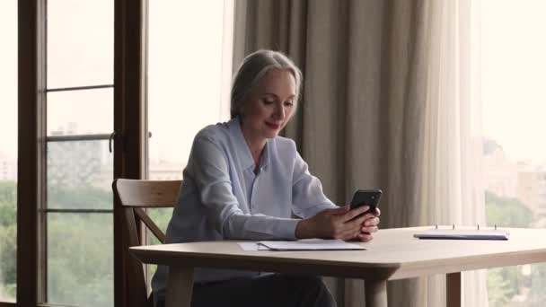 Пожилая женщина сидит за столом с помощью смартфонов онлайн-сервисов — стоковое видео