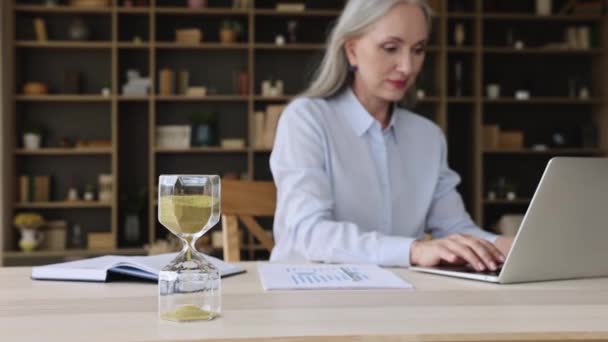 Oudere zakenvrouw werkt op laptop, focus op zandloper — Stockvideo