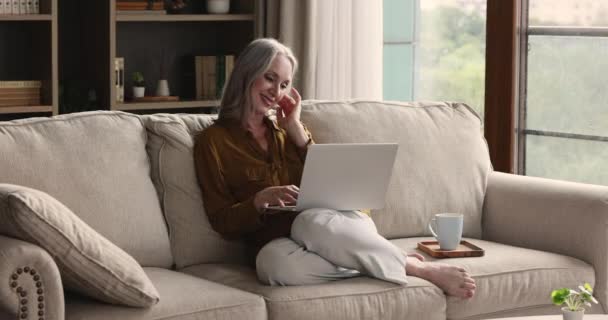 Gammal kvinna sitter på soffan läsa goda nyheter på laptop — Stockvideo