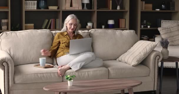 Красива літня жінка розслабляється на дивані з ноутбуком — стокове відео