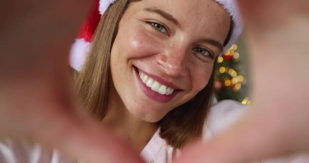 Jovencita celebrar la Navidad mira a la cámara a través del corazón dedo — Vídeos de Stock