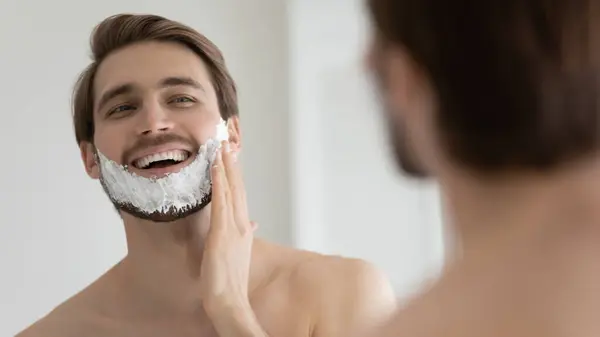 Hombre aplicando espuma de afeitar en la cara delante del espejo —  Fotos de Stock