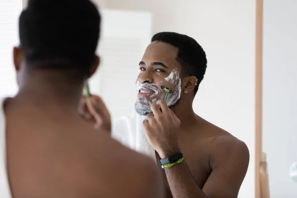 African guy looking in mirror shaving his face with razor — Stock Photo, Image