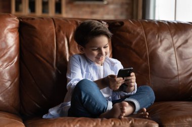 Gülümseyen küçük çocuk akıllı telefon kullanıyor, evdeki koltukta oturuyor.