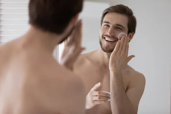 Handsome young man looks in mirror apply facial cream — Stock Photo, Image