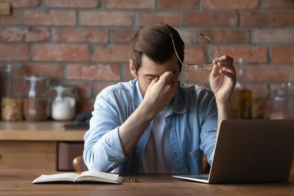 Hombre cansado sosteniendo gafas, frotando los ojos secos, sentado a la mesa — Foto de Stock