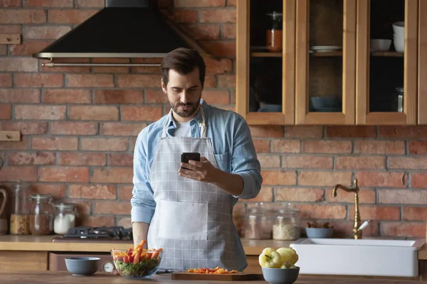 Bearded man using smartphone, distracted from cooking in kitchen — Stock Photo, Image