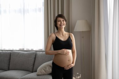 Genç, güler yüzlü, hamile bir kadının portresi ellerini karnına koyuyor.