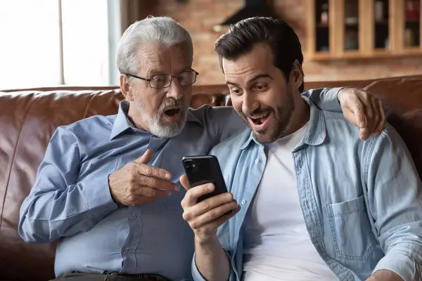 Zdziwiony zaskoczony dwoma pokoleniami mężczyzn patrzących na ekran telefonu. — Zdjęcie stockowe