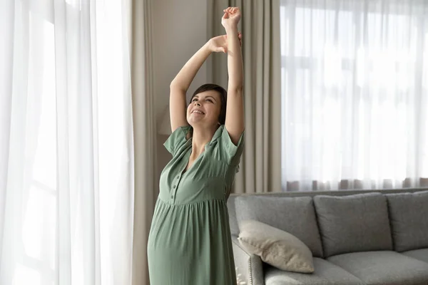 Young future mother stand by window stretch muscles of arms — Stock Photo, Image