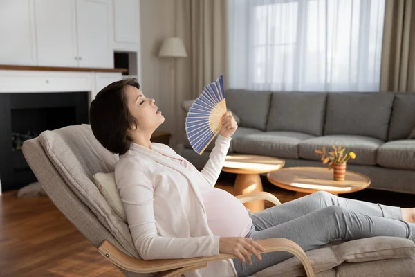 Pregnant female sit on chair cool herself using hand fan — Stock Photo, Image