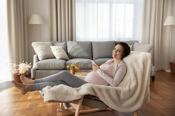 Drømmende, gravid kvinne som ligger i en lenestol og planlegger å kjøpe telefon – stockfoto