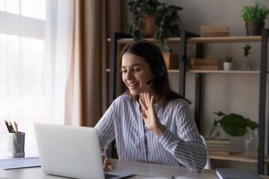 Kulaklıklı gülümseyen kadın el sallıyor, dizüstü bilgisayar kullanıyor, internette sohbet ediyor.