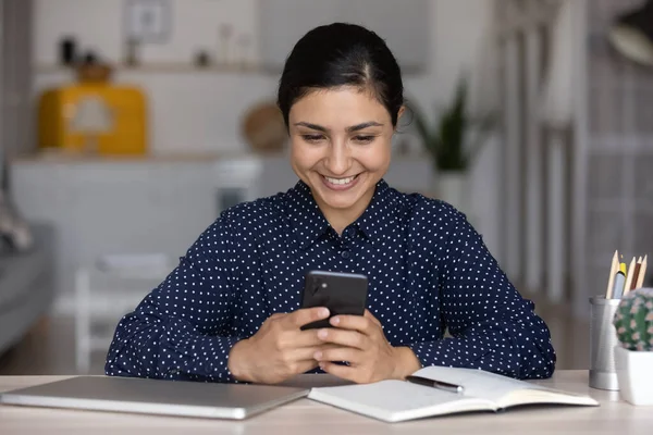 Улыбающаяся индианка с помощью смартфона, сидящая за рабочим столом — стоковое фото
