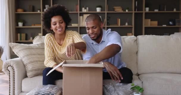 Junges afrikanisches Paar sitzt auf Sofa und packt Paketkasten aus — Stockvideo