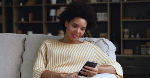 Jovem mulher africana feliz sentar-se no sofá desfrutar usando telefone — Vídeo de Stock