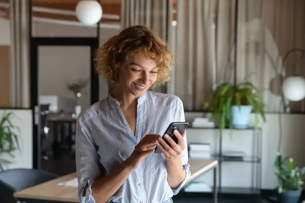 Счастливая молодая самозанятая женщина, использующая сотовый телефон в офисе — стоковое фото
