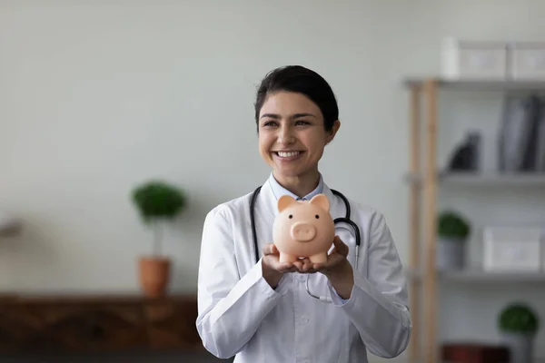 Happy Indian young female doctor showing piggy bank at camera