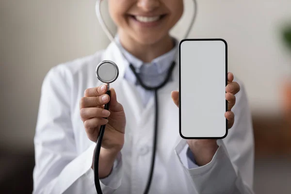 Happy female doctor holding stethoscope, showing blank smartphone screen