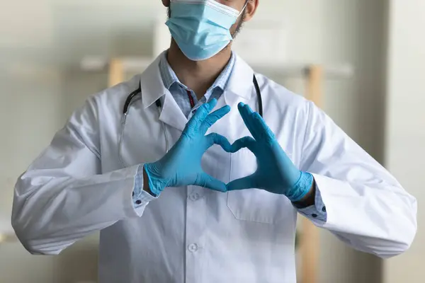 Joven doctor en mascarilla facial, guantes de látex mostrando el corazón del dedo —  Fotos de Stock