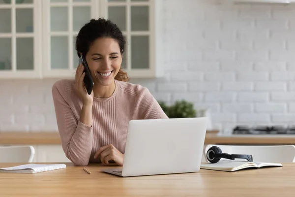 Smiling young woman work online on laptop talking on cellphone — Stock Photo, Image